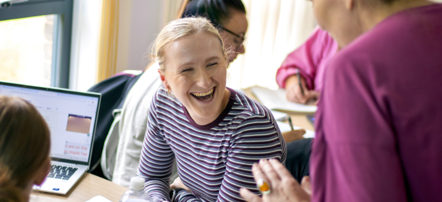 An apprentice sat at a desk, smiling up at a lecturer with other apprentices working around them